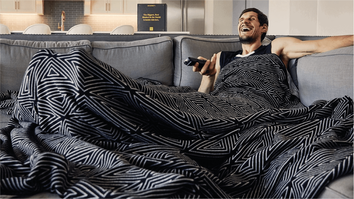a guy laughing with a Tv remote in his hand as he is cozed with a big blanket. 