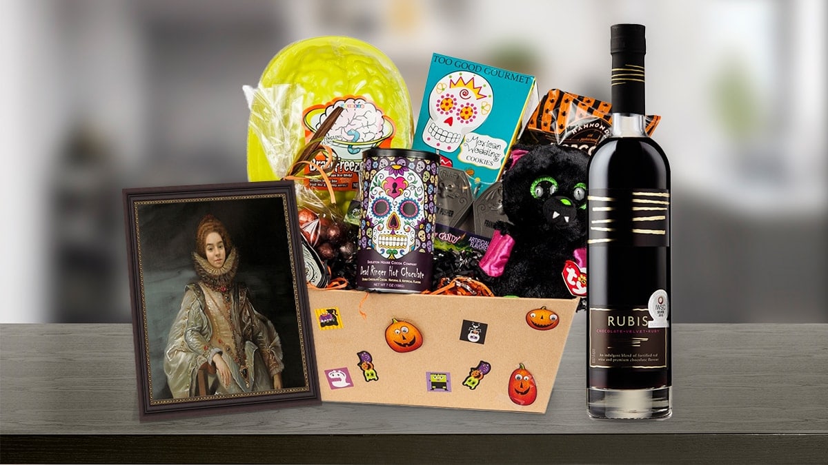 A Halloween gift basket kept on a table containing, wine, candies, cookies, chocolates, and a framed painting of a woman, that can be given as a Halloween gift.