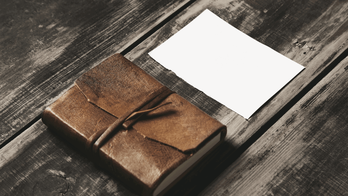 A brown leather journal kept on a wooden surface with a white page nest to it. 