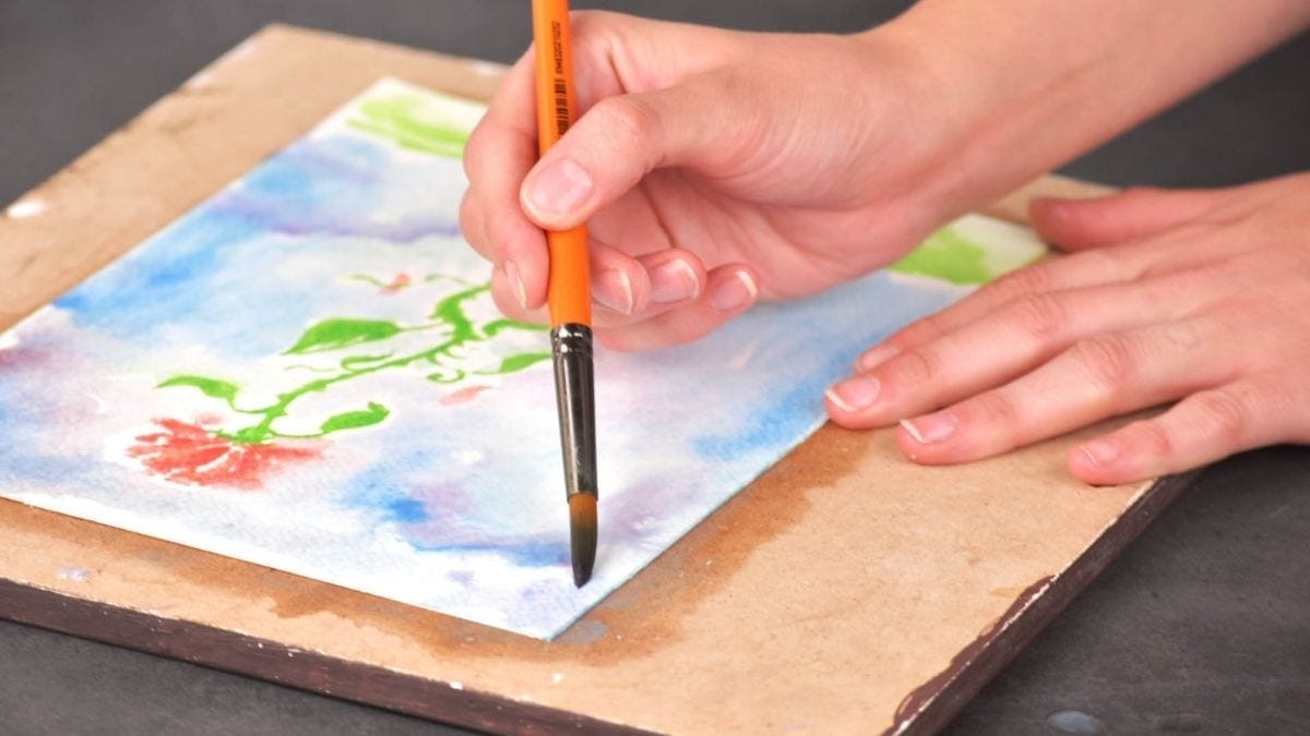 A hand holding a soaked paint brush and painting watercolor on a cubicle wooden board. 
