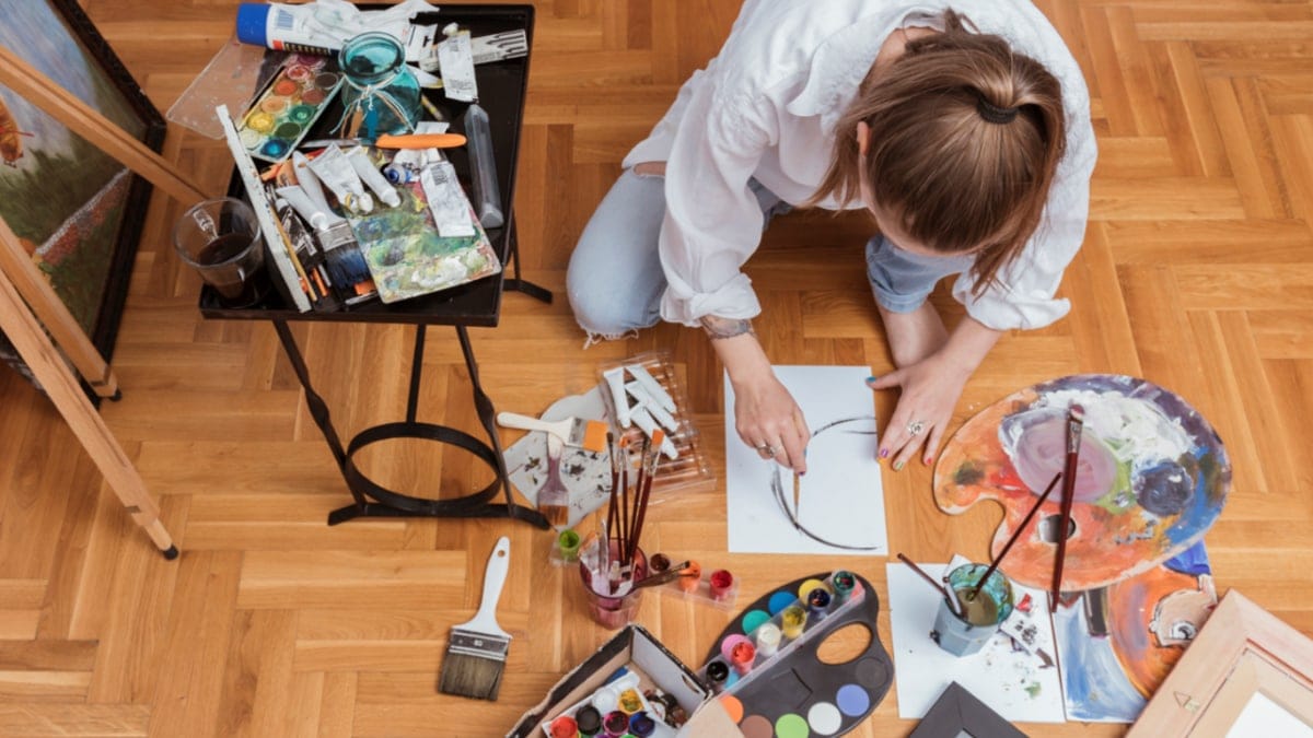 A woman sitting on the floor with acrylic painting supplies around her and she is painting on an empty canvas with a brush as one of the acrylic painting tips for beginners.