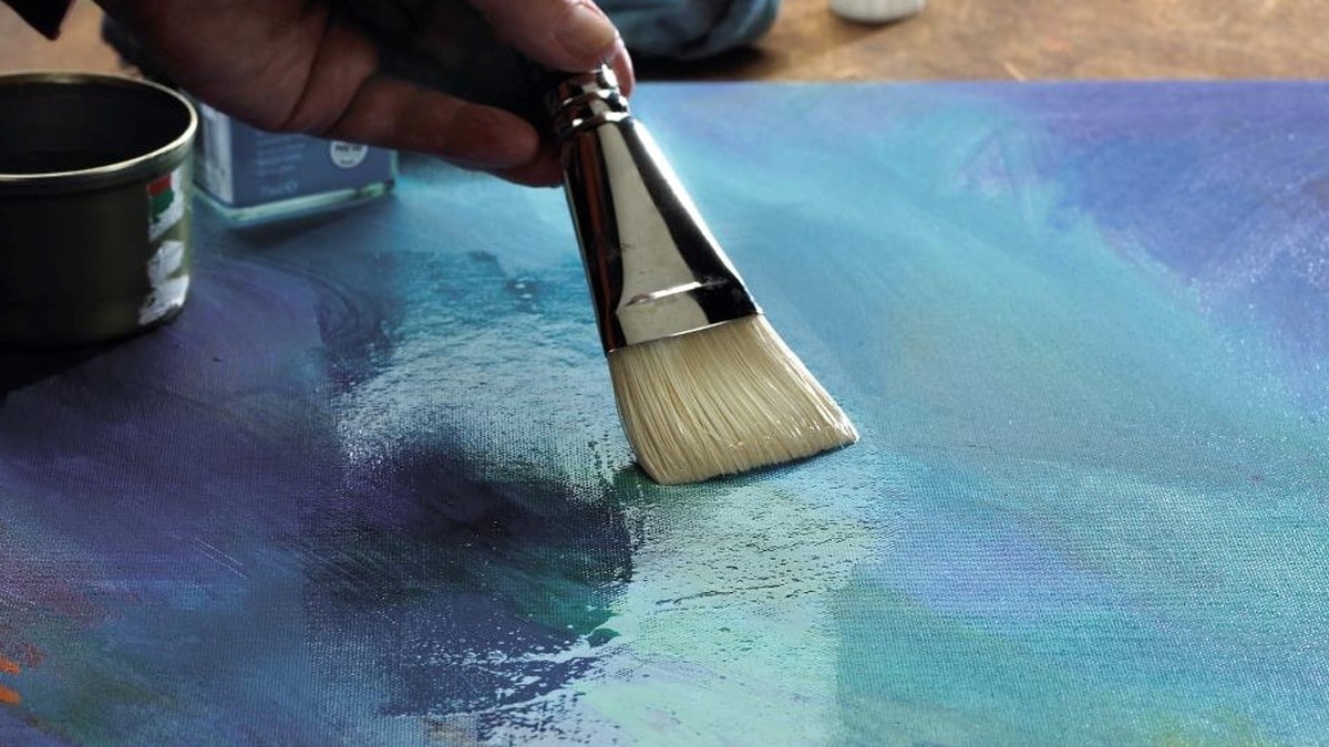 a hand applying clear varnish with a paint brush on a painted canvas.