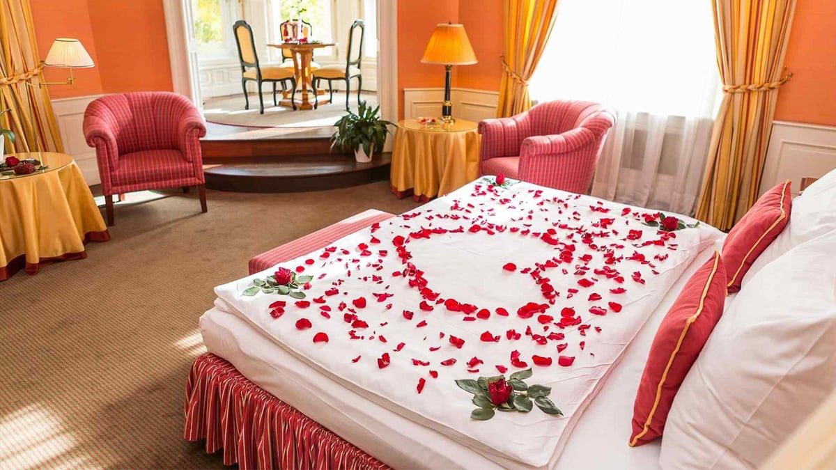 A bed is decorated with rose petals to make a heart. the whole room decor is complimented by orange-red-pink-white color scheme.