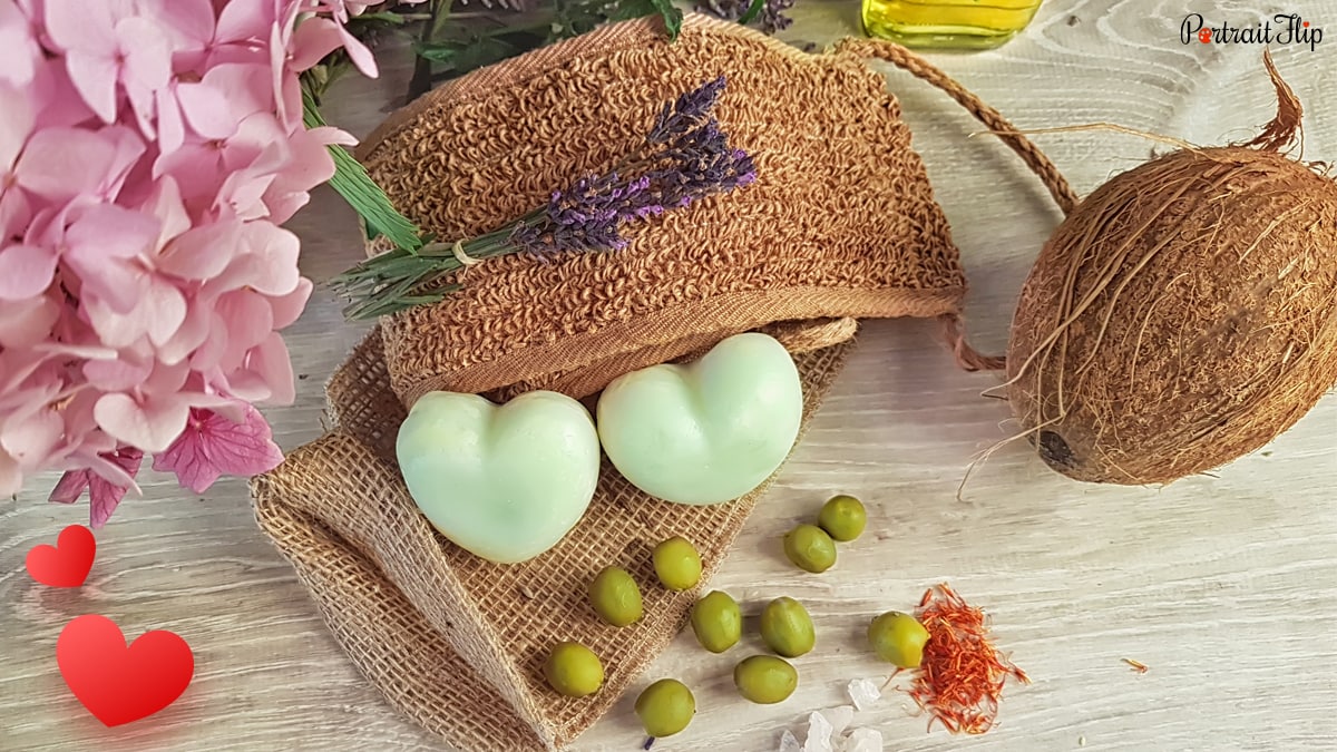 green colored heart soaps is kept beside the ingredients like olives, coconut, flower, saffron 