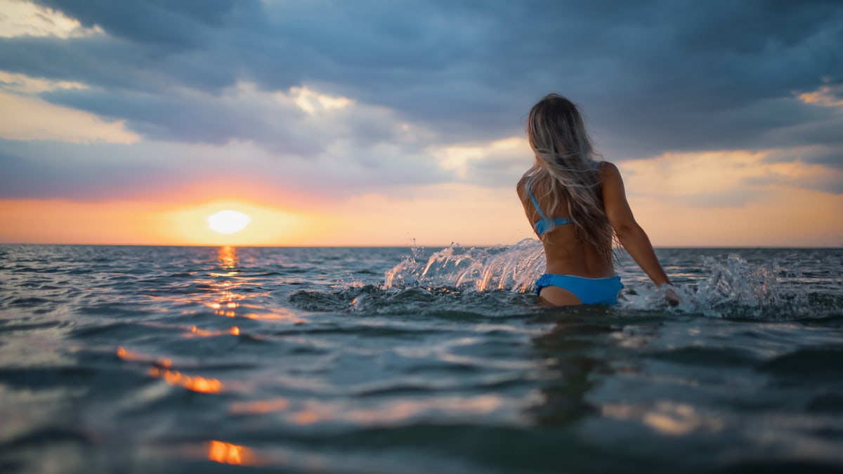 a woman in a bikini splashing water in the sea while looking at the setting sun. 