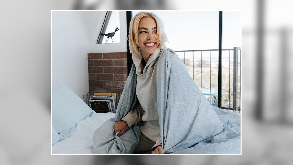 a smiling woman with a gravity blanket wrapped around her on bed. 