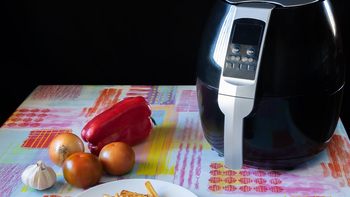 a black Air fryer on the table. beside it, is red capsicum, onions, and garlic. 