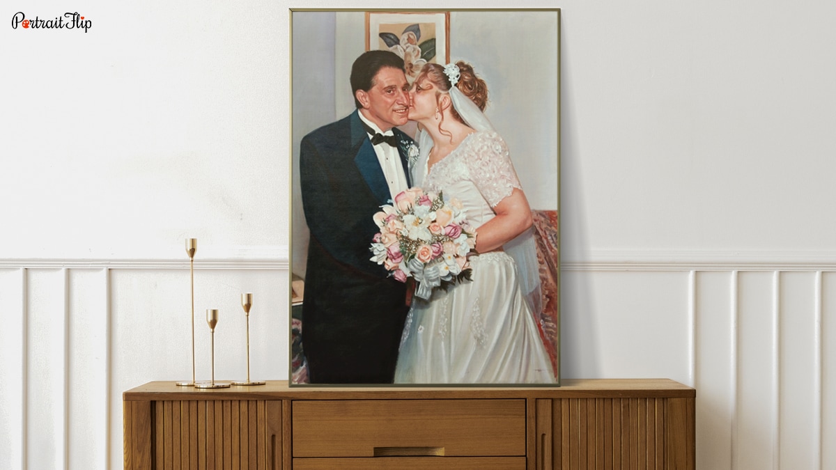 A handmade custom portrait of two people on their wedding day, the bride is kissing the grooms cheek. This portrait is on a table with decorative pieces around the painting.