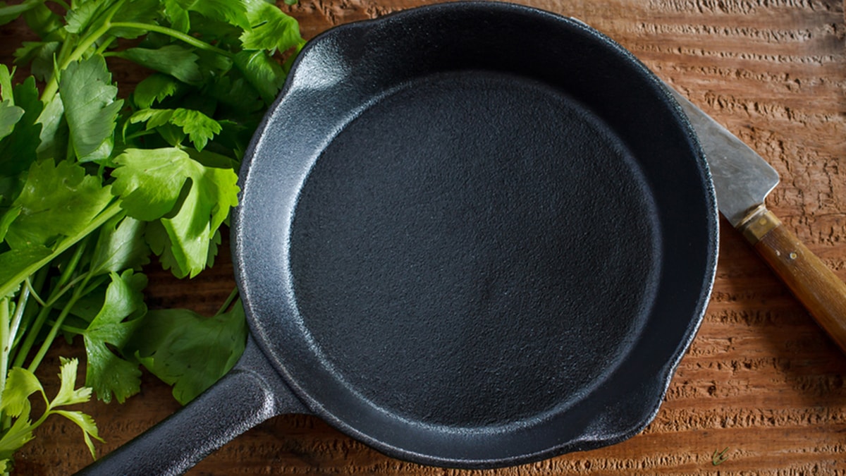 An iron skillet sitting on a kitchen counter with some coriander on the side and a knife. The iron skillet is one of the graduation gift ideas for sister.