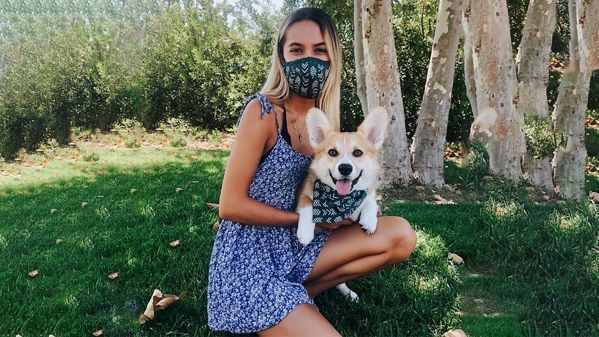 A lady and her dog sitting down in the grass while the lady holds her dog and they are wearing matching mask and bandana set.