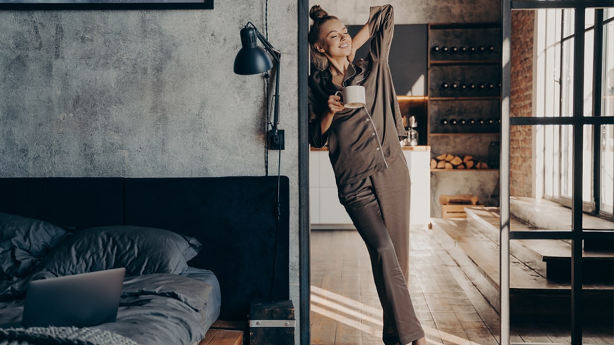 A girl relaxing in a satin pajama set with a cup of coffee in her hand inside her bedroom