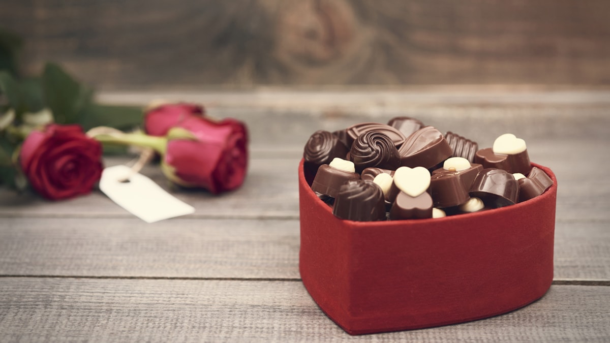 A couple of roses and a heart shaped box in red color filled with different chocolate variants.