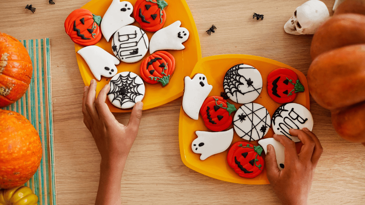 A plate full of Halloween cookies displayed on a Halloween themed table with pumpkins, skull and spiders around the plates. two hands reaching out for the cookies and grabbing them.