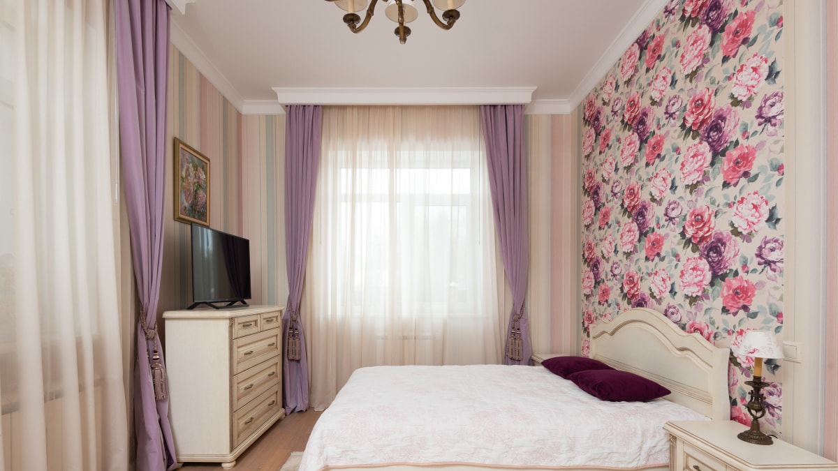 A floral wallpaper displayed on a bedroom wall with purple and white curtains and a white bed in the middle.
