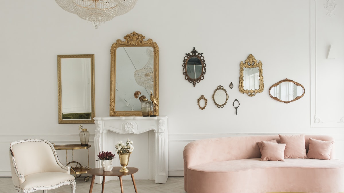 Different antique frames placed on a living room wall with a pink couch and wooden table and a white chair in the living room.
