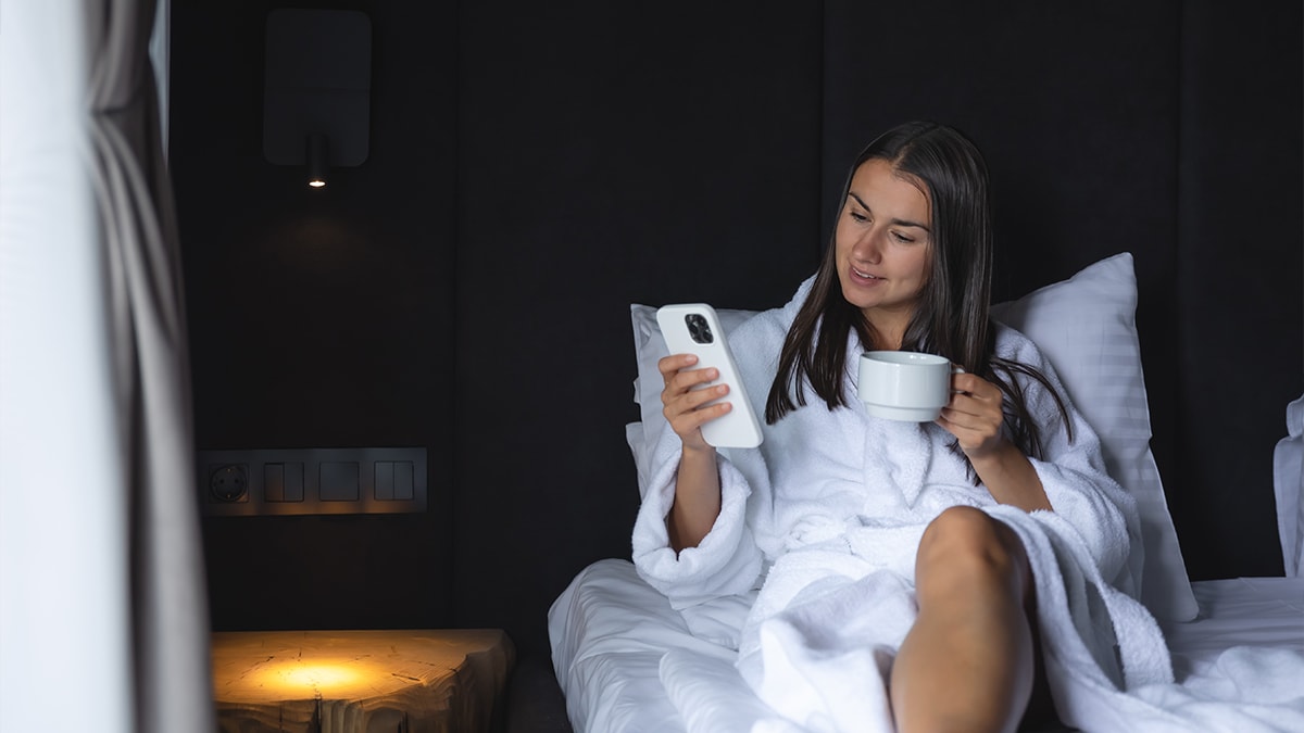 A Woman wearing a white bath robe while leaning backwards on bed with phone and cup in hand.