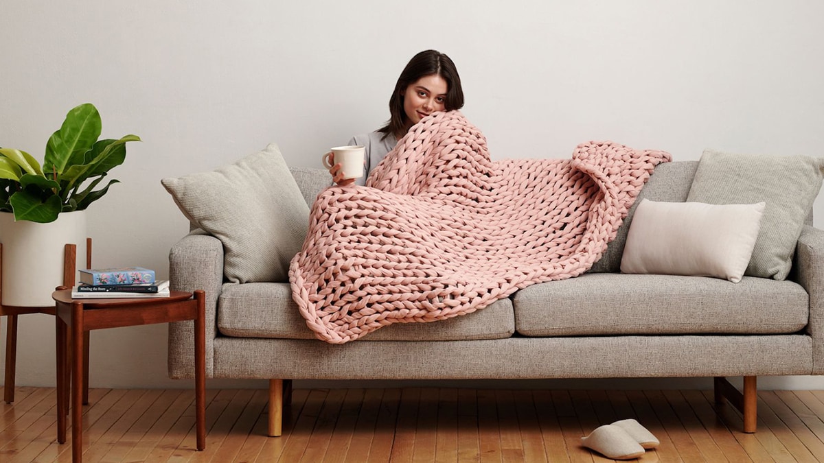 A woman sits on a sofa in the living room while holding a cup and snugging a Bearaby Cotton Napper.