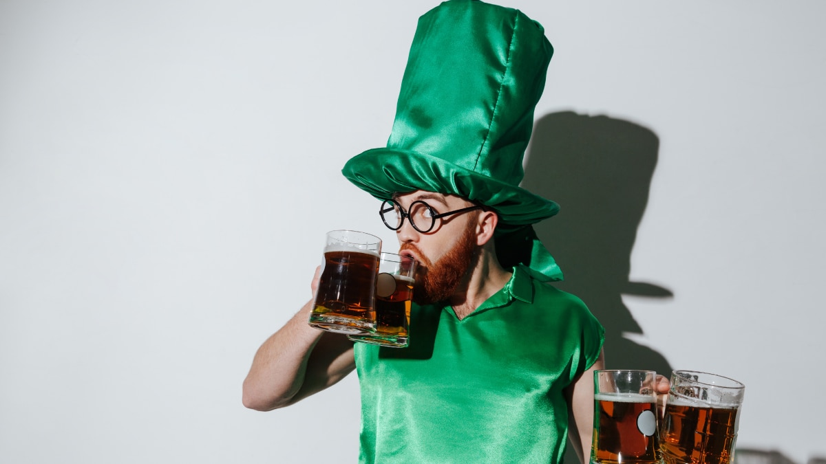 A man wearing green satin cloth and hat with four beer glasses in hand while drinking