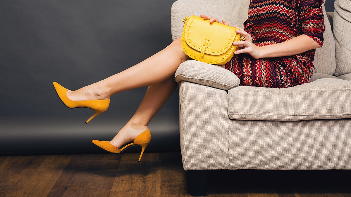 A woman's legs are displayed as she reclines on a couch wearing heels and a yellow bag.