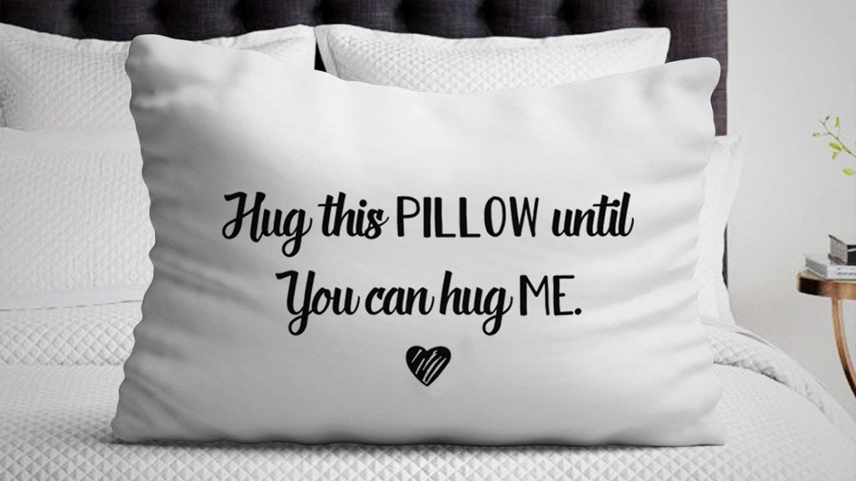 A customized white pillow with the black text Hug this pillow until you can hug me including a heart print
