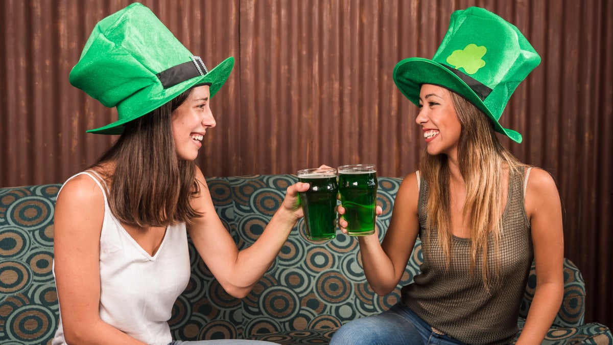 Two woman cheering with green drink glasses wearing a big green hat while sitting on a sofa