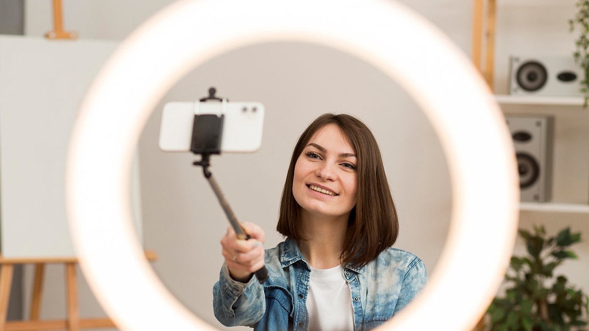 A woman takes a selfie with a selfie stick while being framed by a vlogging ring light.