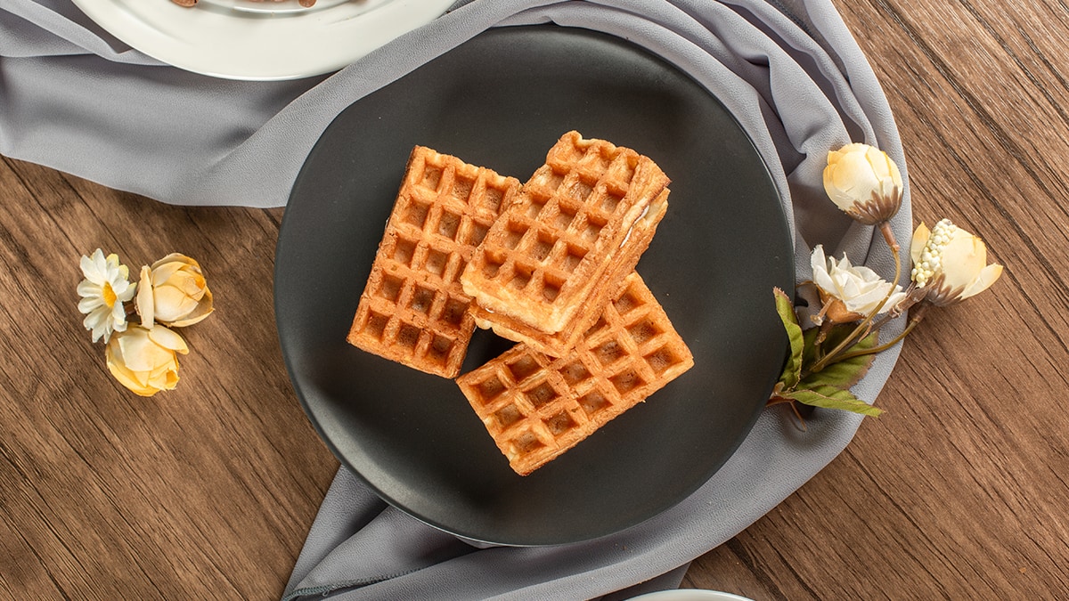 On a wooden table, there is a black dish with waffles and white artificial flowers wrapped around a grey cloth.