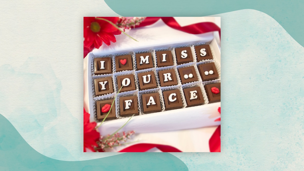 18 small pieces of chocolate with letters and heart shapes on them kept in a white colored box alongside red ribbons and red flowers. 