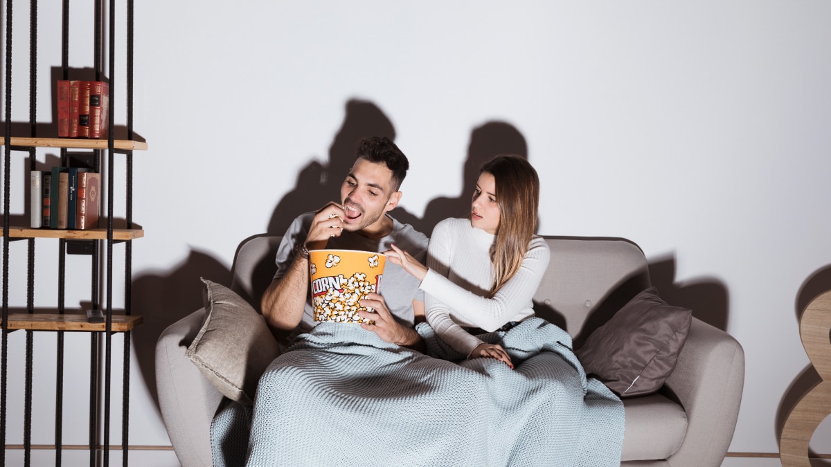 A couple sitting on a couch with blankets on while the man is having a popcorn 