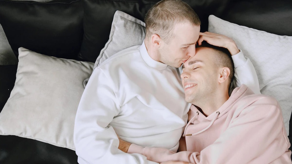 A gay man kissing another on his forehead while lying on the bed.