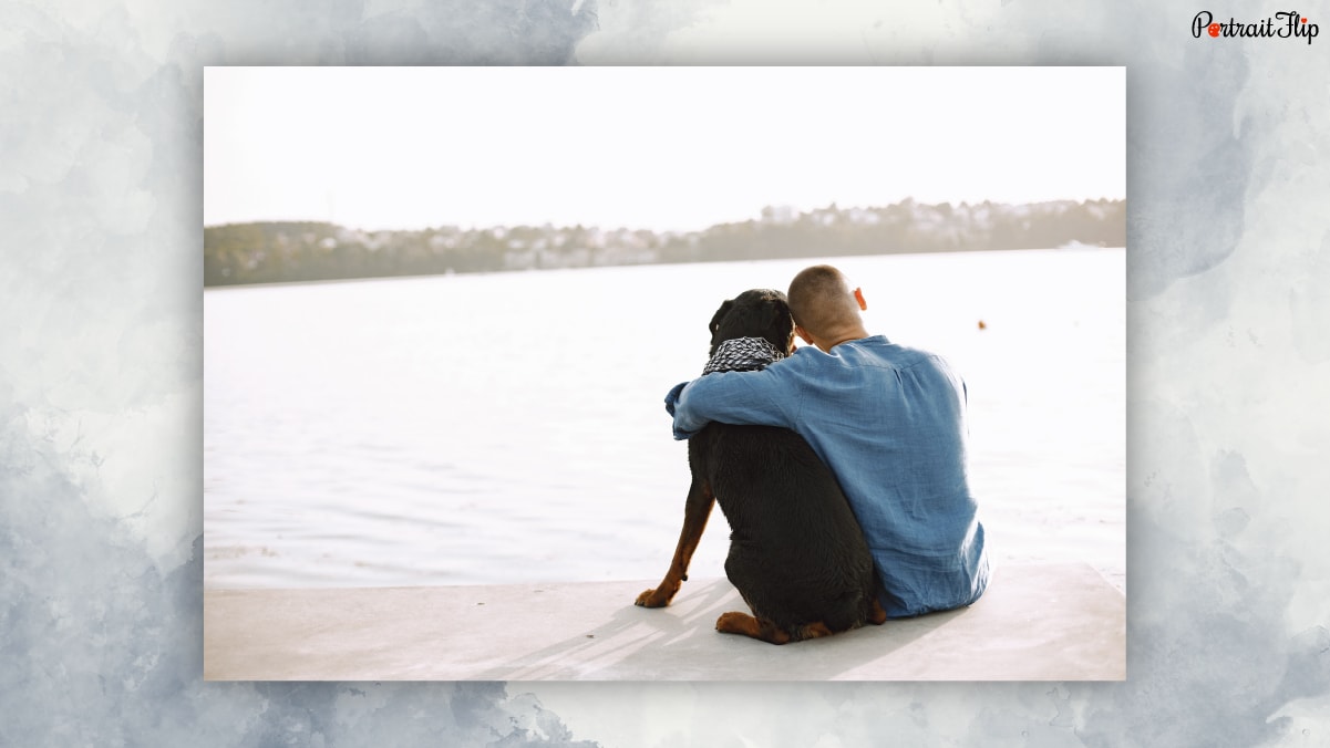 Behind view of a man giving a side hug to the dog sitting in front of a sea view. 