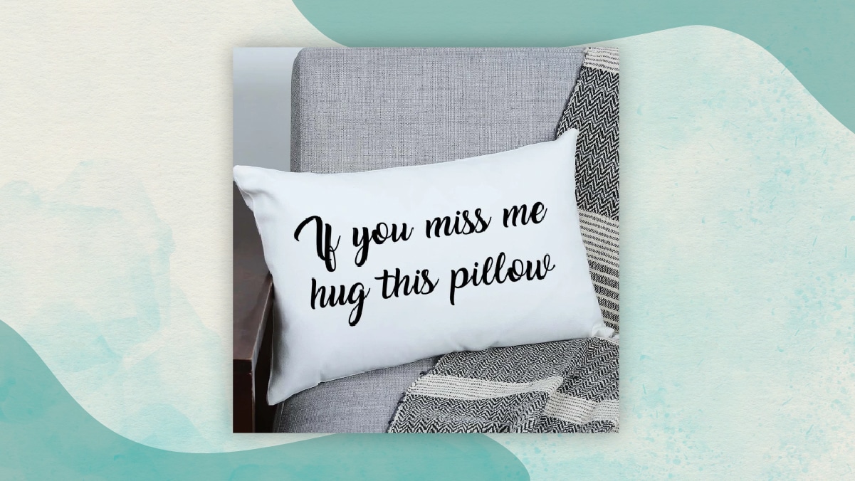 A white colored pillow with" If you miss me hug this pillow" written on it with black ink kept on a grey sofa. 