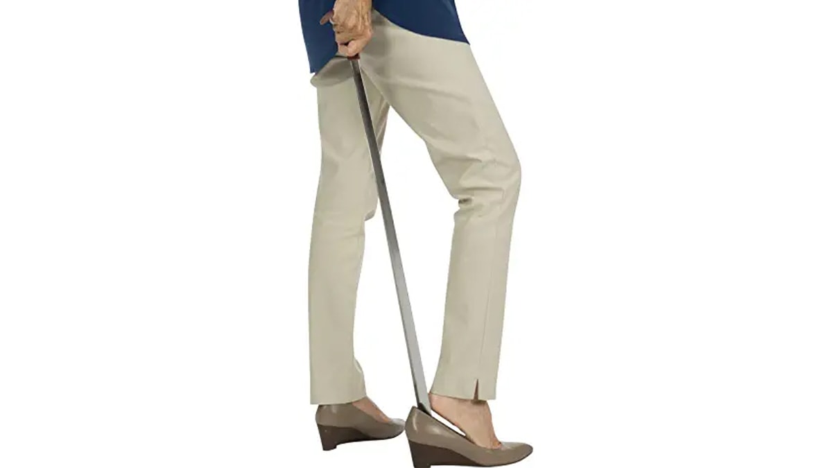 A women wearing brown pants holding a shoe horn to wear shoes in a white background. 