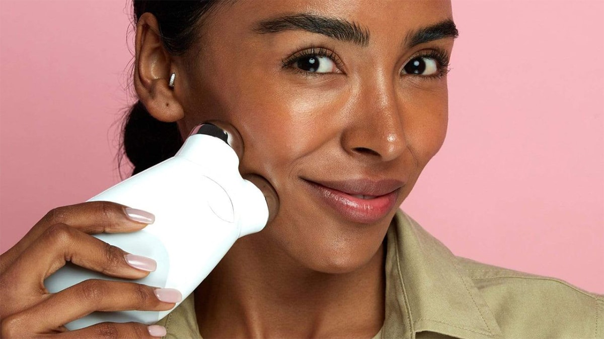 A smiling women using a skincare product as mother's dat gift idea on her face in a pink background. 