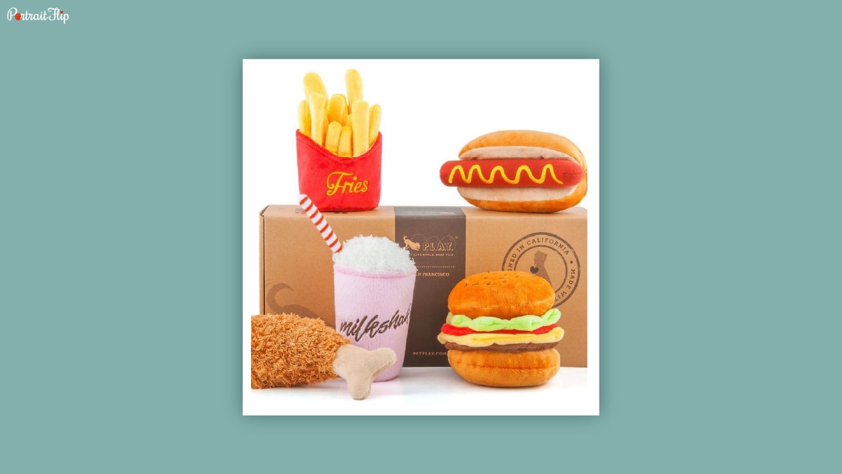 A set of soft toys shaped like burger, fries, chicken leg piece and milkshake kept on a white background as dog birthday gifts. 