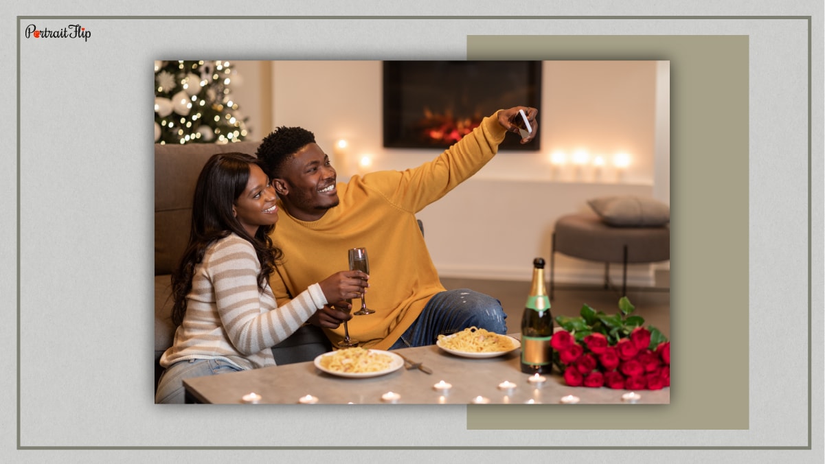 A couple sitting next to a couch with glass of wine in their hand and on the table is two plates of pasta, roses, mini candles and a bottle of wine. 