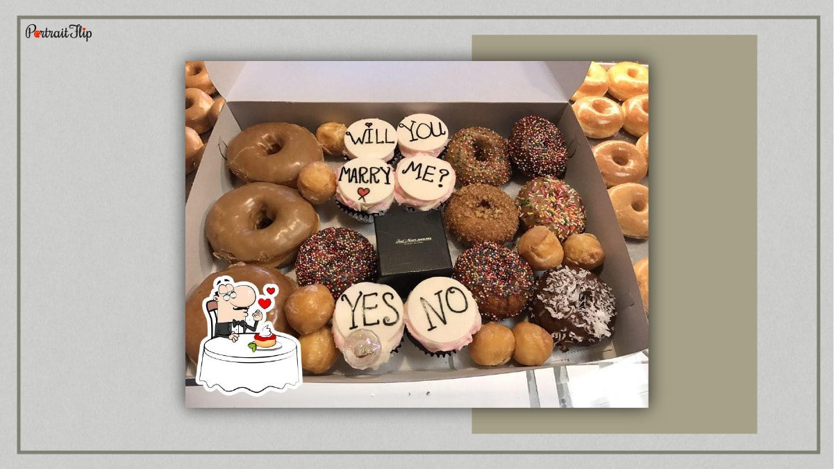 A box full of donuts with a box of ring in between and few cupcake customized with a special frosting of a proposal. Written over the cupcake is will you marry me? Yes or No