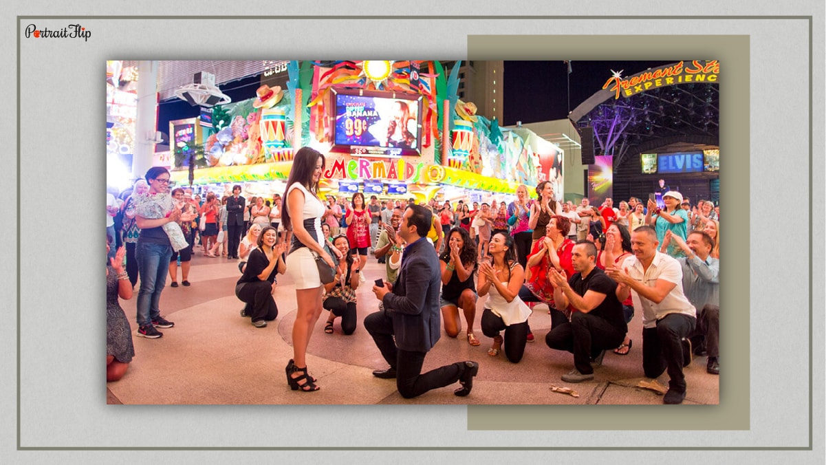 In this image the man came up with a unique proposal idea. He proposed his woman on a square with a flash mob performance and is now sitting on his knee with a ring in his hand. 