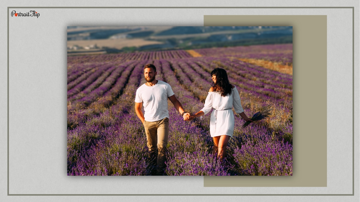 A couple amid a lavender field. Both are wearing white clothes and are holding hands and walking past the field. 