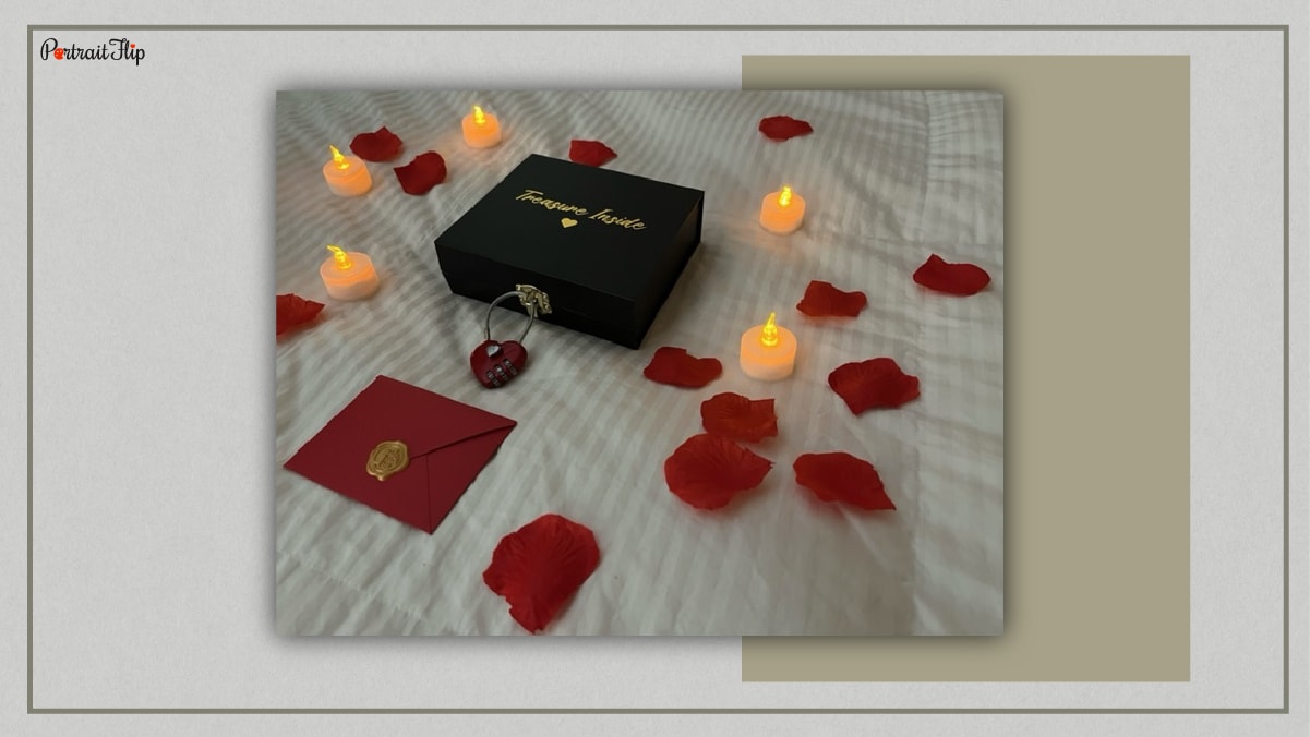 A treasure hunt black coloured box which is locked with a heart shaped lock and is kept on a mattress surrounded with electric candles and rose petals and also a red coloured envelope is kept on the front of the box.