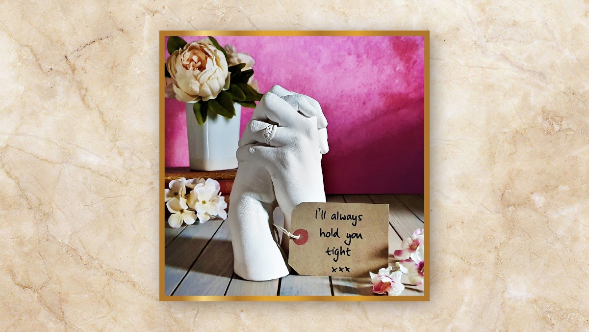A holding hands sculpture kept on wooden table with a pink wall behind it along with white colored flowers beside.  it as personalized wedding gifts.  