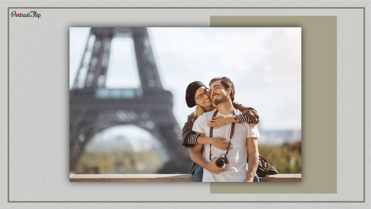 A couple snuggling in front of the Eiffel Tower and the woman is hugging the man from the back while the man is holding his camera.