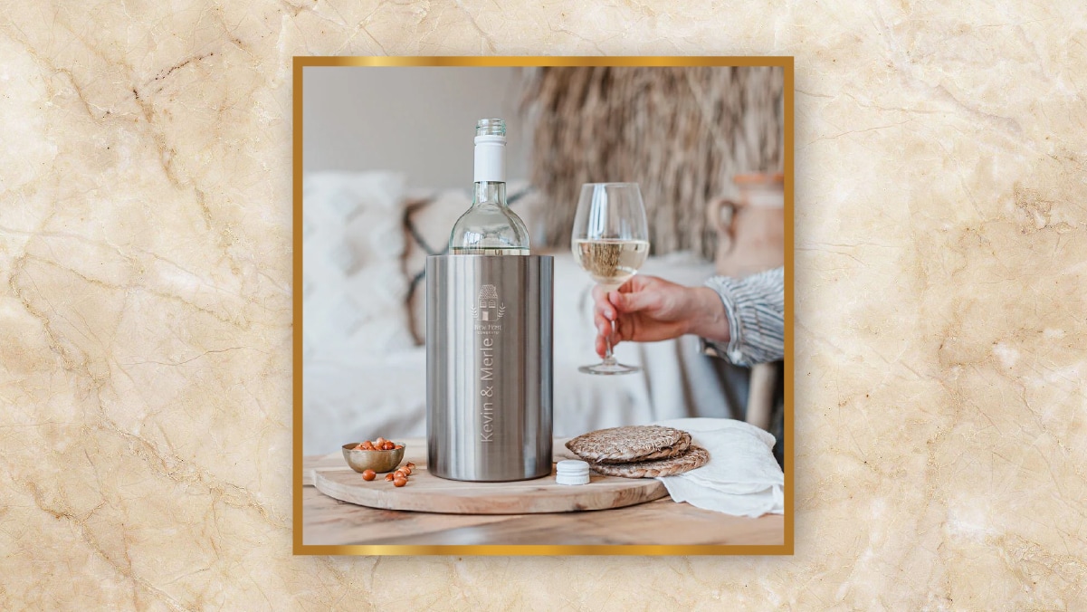 A silver colored wine chiller as a personalized wedding gift with a bottle of wine in it kept on a wooden table with a hand holding a wine glass with wine in it beside it. 