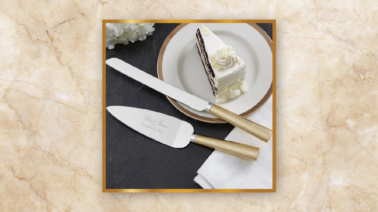A pair of engraved knife with golden handles kept beside a piece of cake in a white dish on a dark brown table as personalized wedding gifts. 