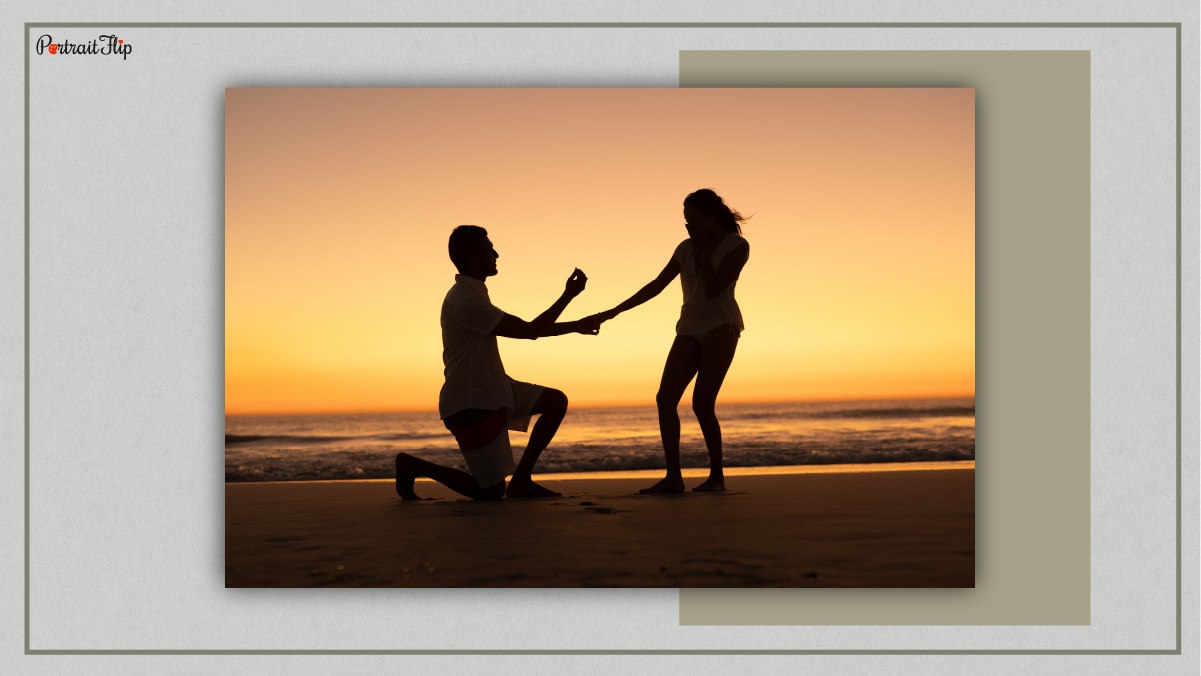 A man on his knees with a ring in his hand proposing to a woman who is surprised. It is all happening during the time of the sunset at a beach.