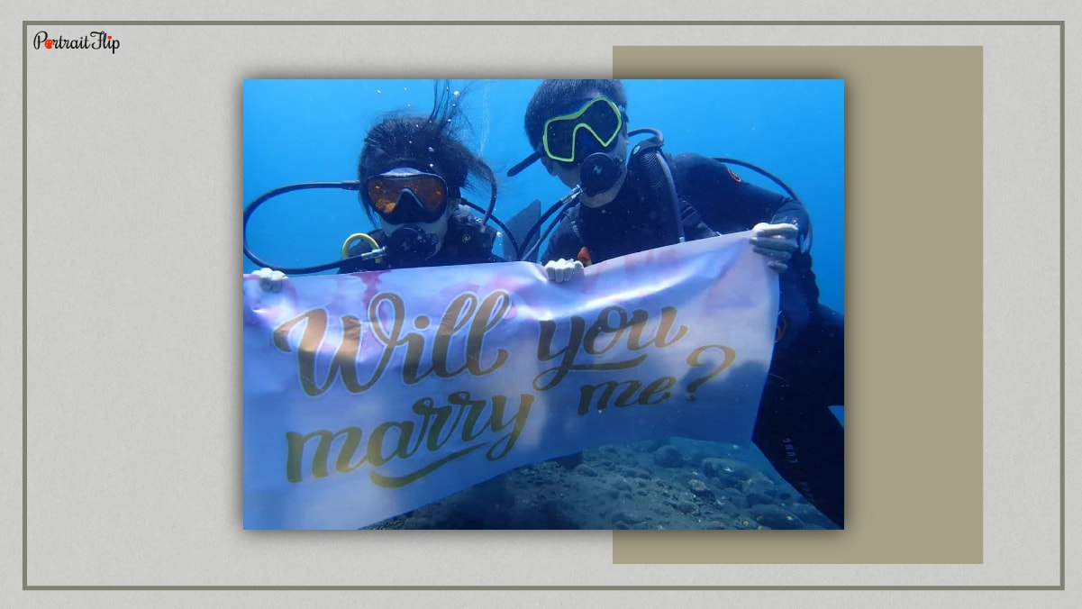 A couple is scuba diving while holding a banner which reads Will you marry me? 