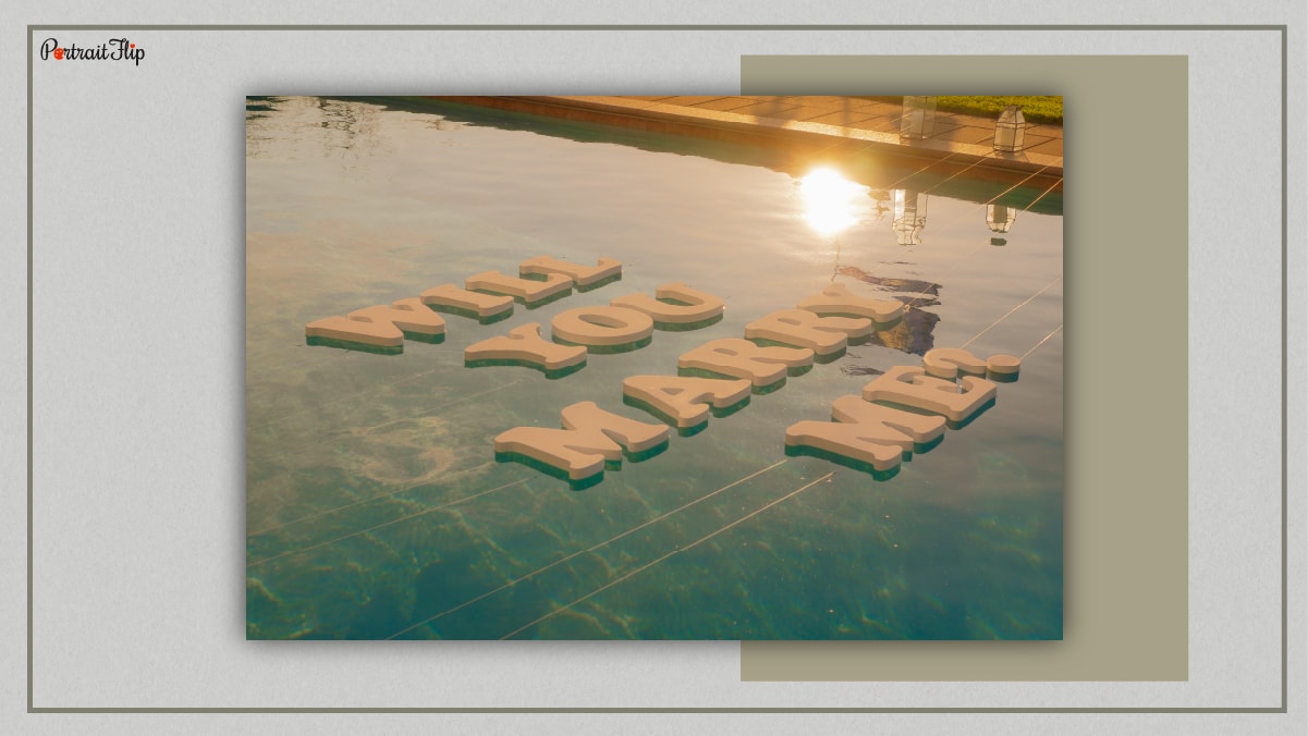 Floating letters over a pool which reads Will You Marry Me? 
A sun can be seen as well in the shadow on the water surface.