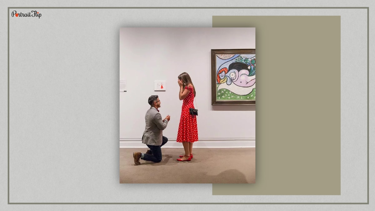 A man sitting on his knees and proposing a woman who is standing in a red dress with both hands on her face. They both are in an art gallery. 