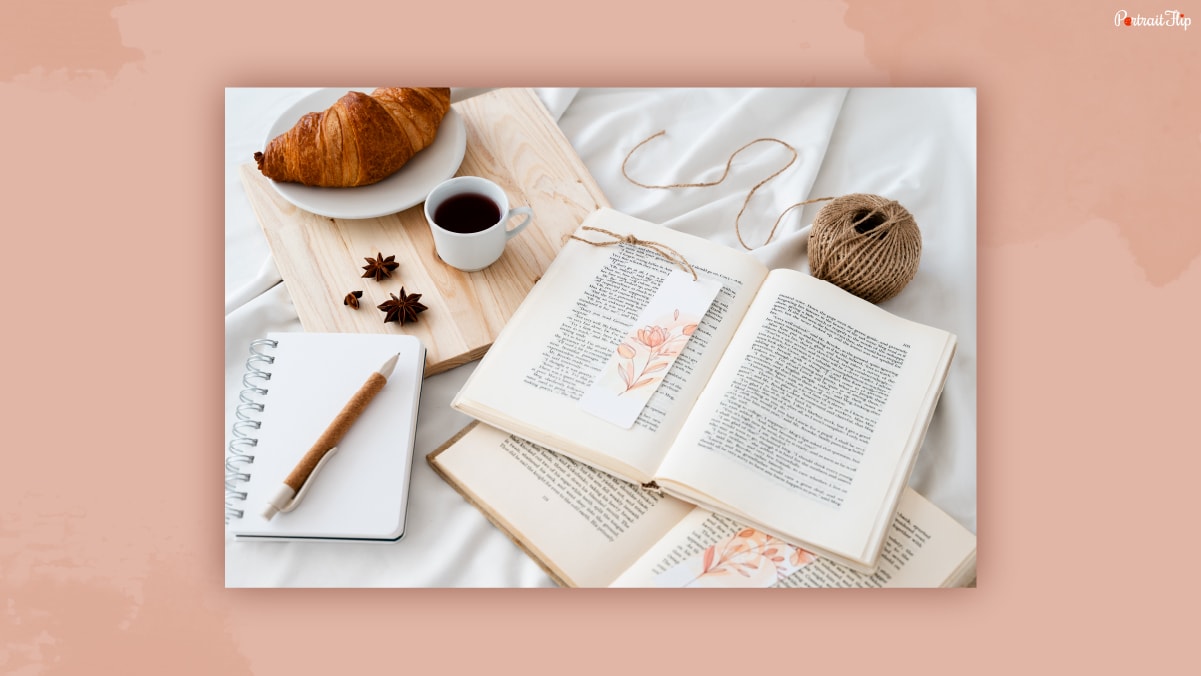 A bookmark kept in between the books beside a blank notebook with croissant, tea and wool on a white bedsheet.
