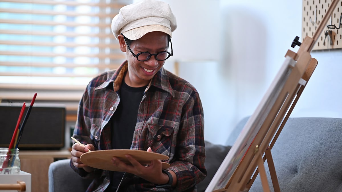 a man is smiling, holding a brush and palette in his hands, looking at the canvas